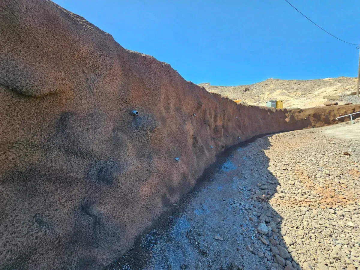 Una pared marrón curva y texturizada corre a lo largo de un camino de grava, con un cielo azul brillante sobre ella.