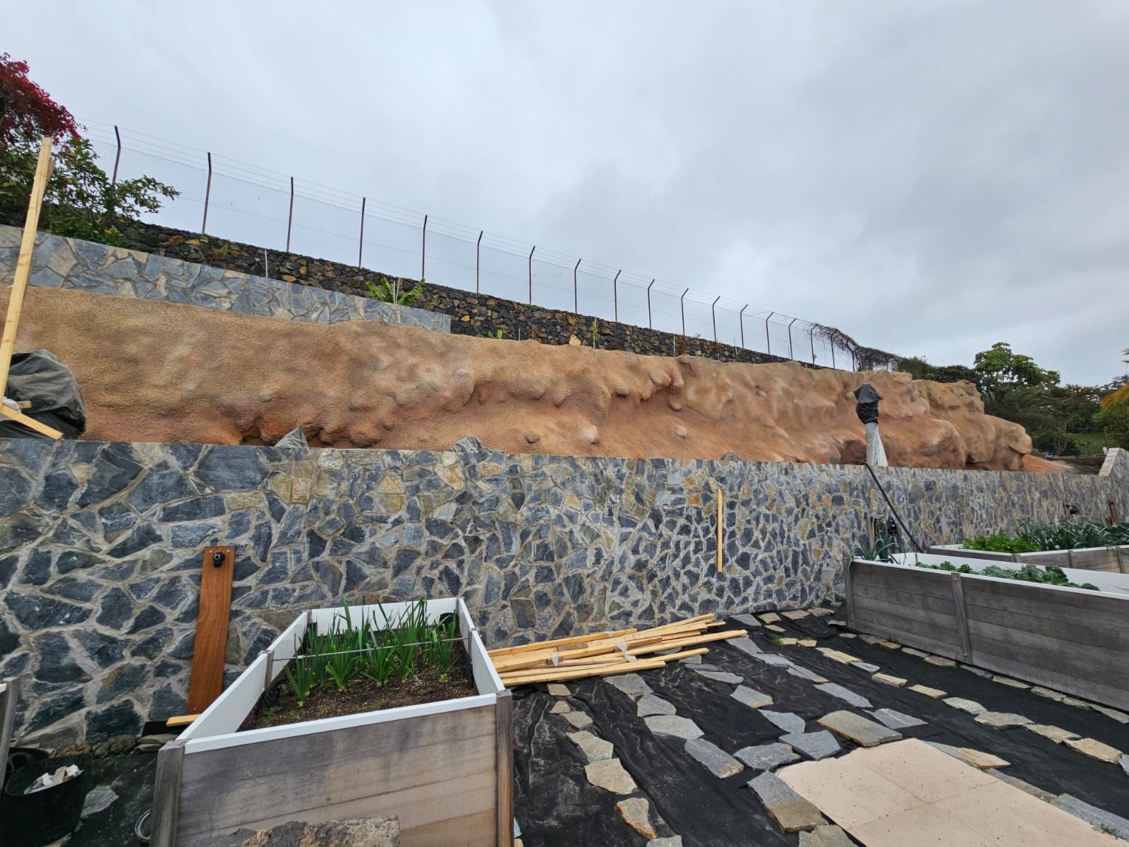 Muro de contención de piedra con parterres escalonados y una persona de pie en un nivel superior, bajo un cielo nublado.