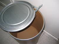 Un bote de basura con tapa de metal se encuentra sobre un piso de baldosas.