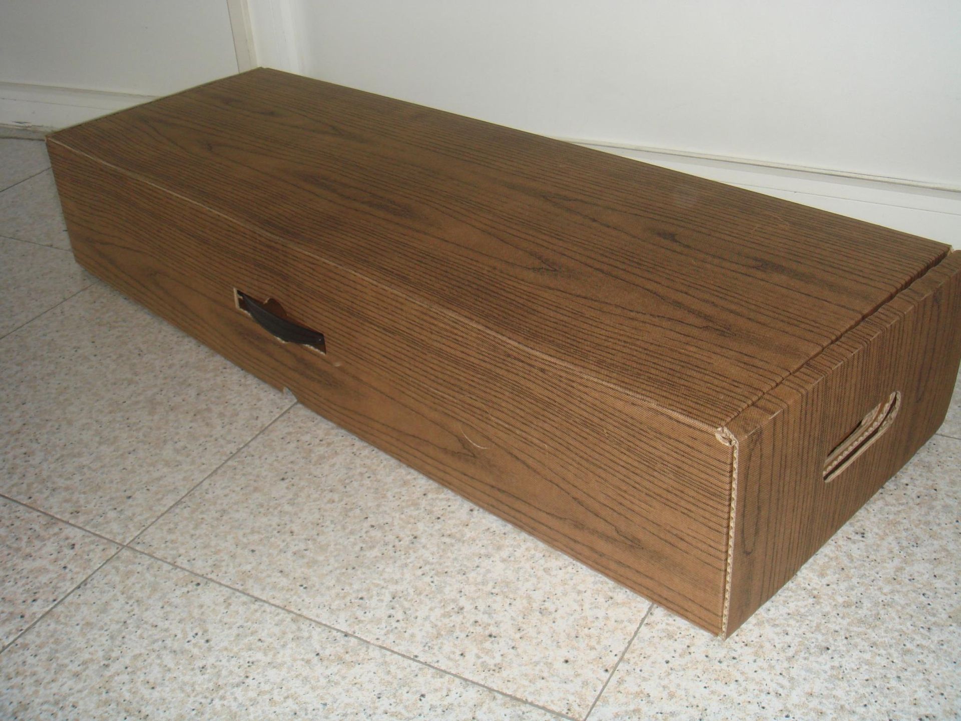 Una caja de madera con asa reposa sobre un suelo de baldosas.