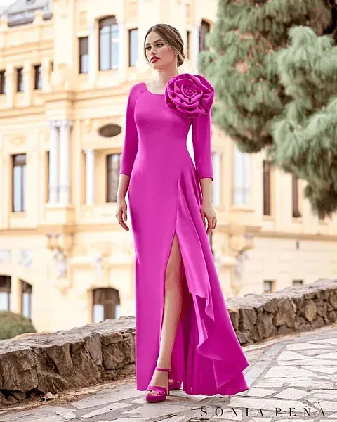 Una mujer lleva un vestido largo rosa con una flor en el hombro.