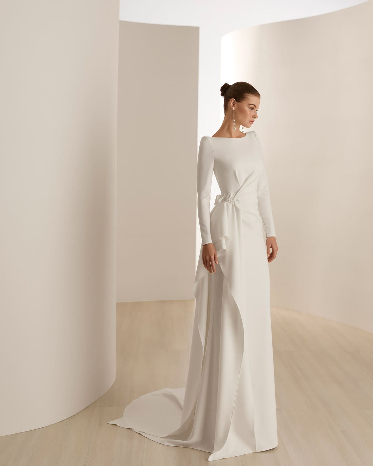Mujer con un vestido blanco largo de pie en una habitación minimalista de color beige con paredes curvas.