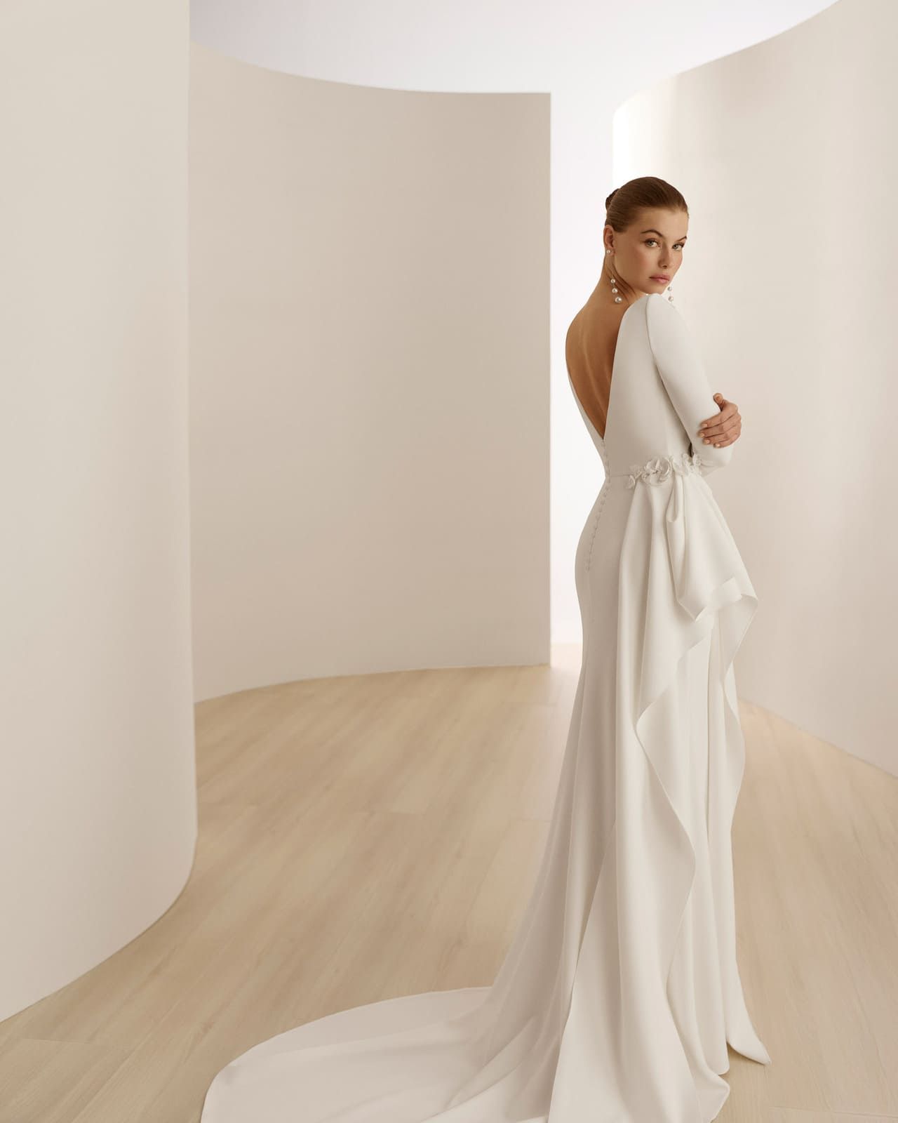 Mujer con vestido blanco de pie en una habitación minimalista de forma curva, mirando por encima del hombro.