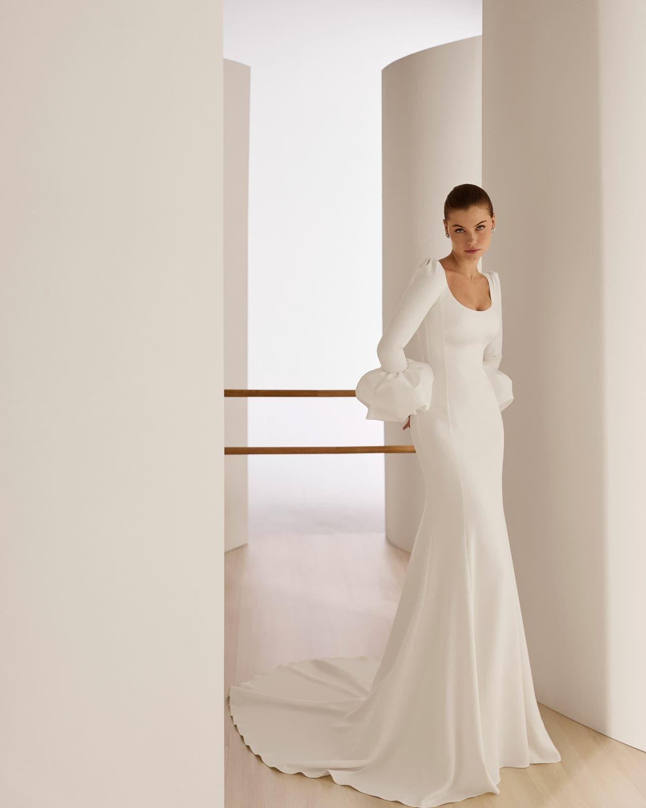 Mujer con un vestido blanco largo de pie en un pasillo moderno sosteniendo un ramo de flores blancas.