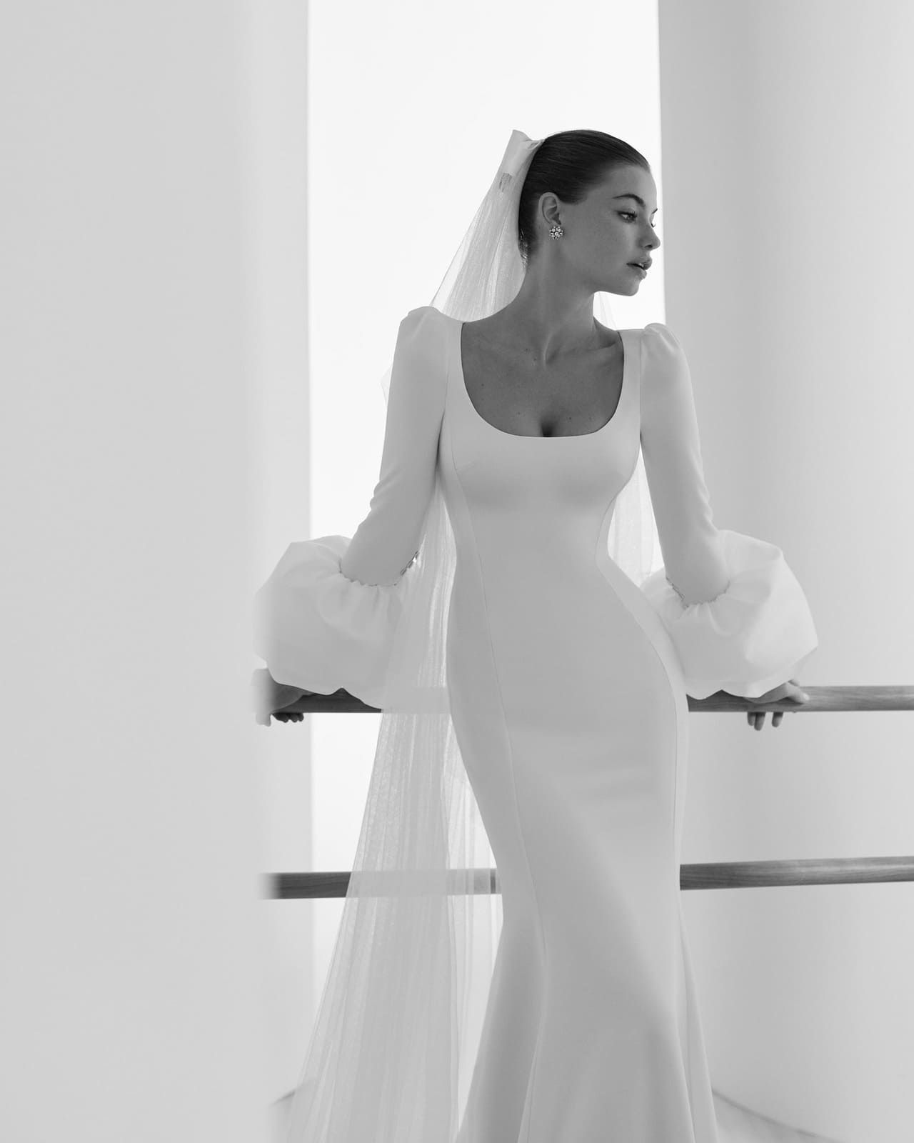 Novia con un largo vestido blanco y velo, de pie junto a una barandilla, mirando hacia un lado.