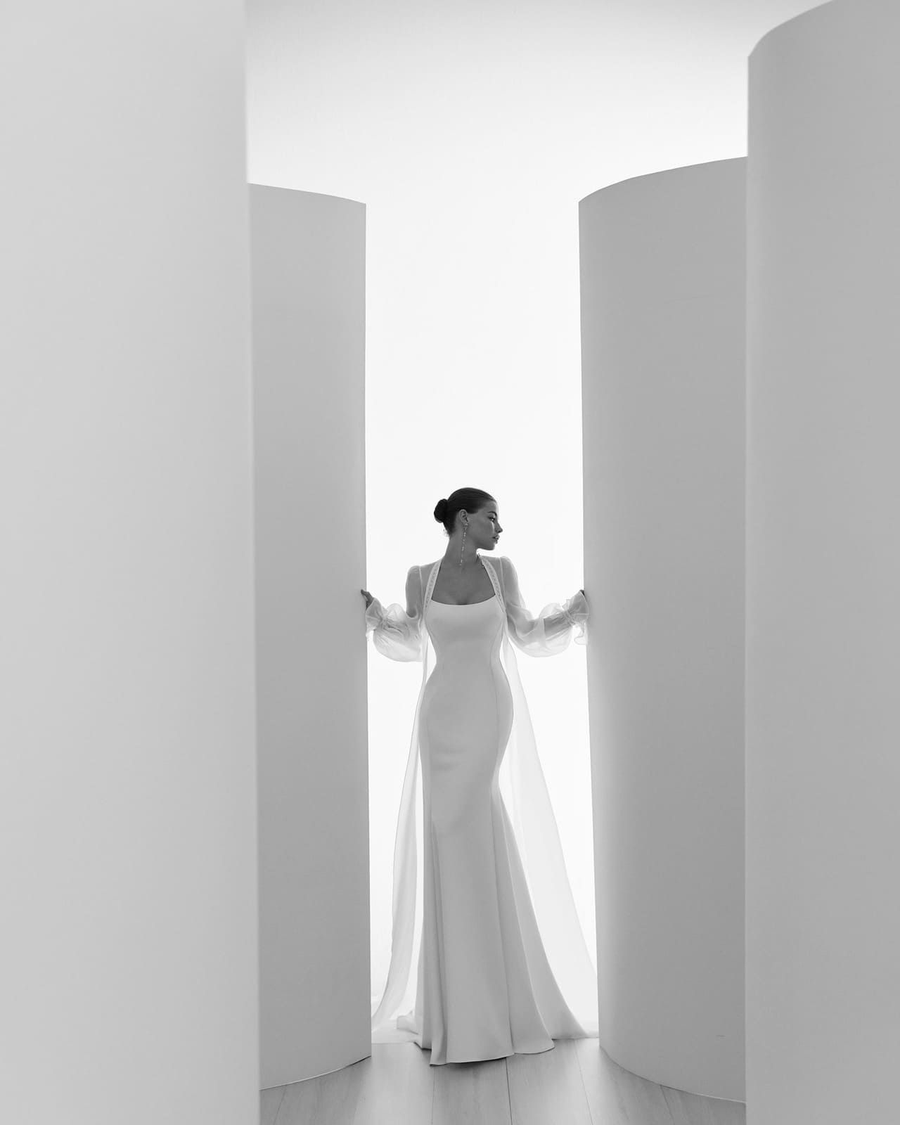 Mujer con vestido blanco de pie entre altas columnas blancas curvas en un espacio luminoso y minimalista.