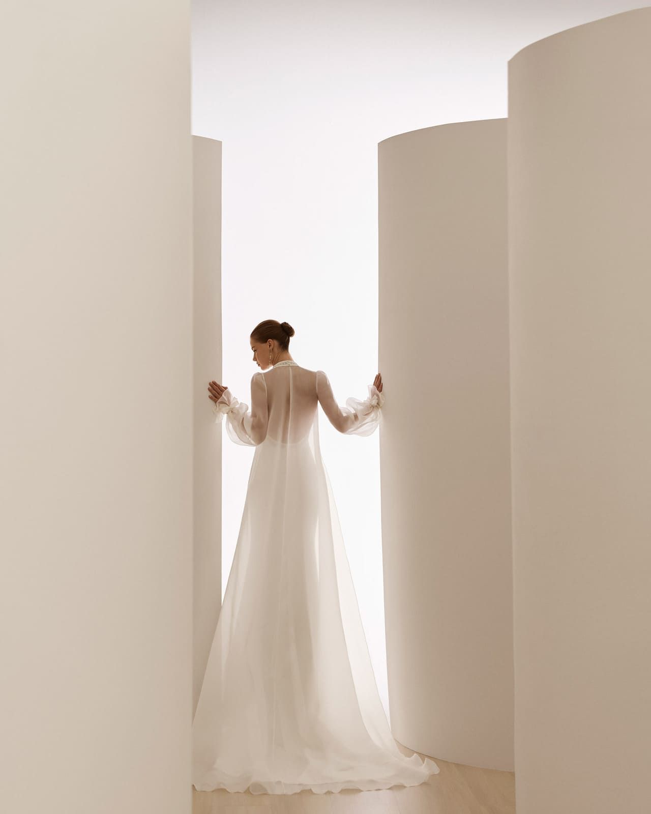 Persona con un vestido blanco largo de pie entre altas paredes blancas curvas en un espacio luminoso y minimalista.
