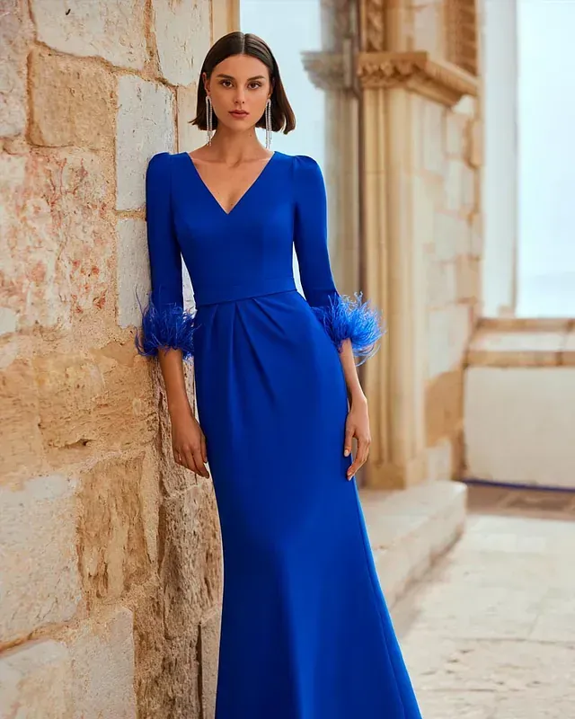 Una mujer con un vestido largo azul con plumas en las mangas está apoyada contra una pared de piedra.