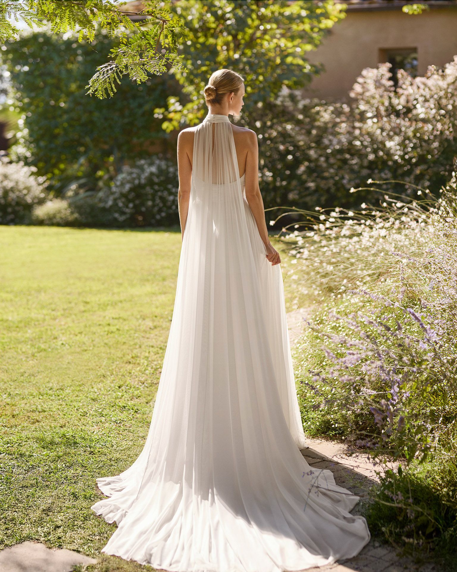 Mujer con un vaporoso vestido blanco de pie en un sendero soleado de un jardín, junto a flores y vegetación.