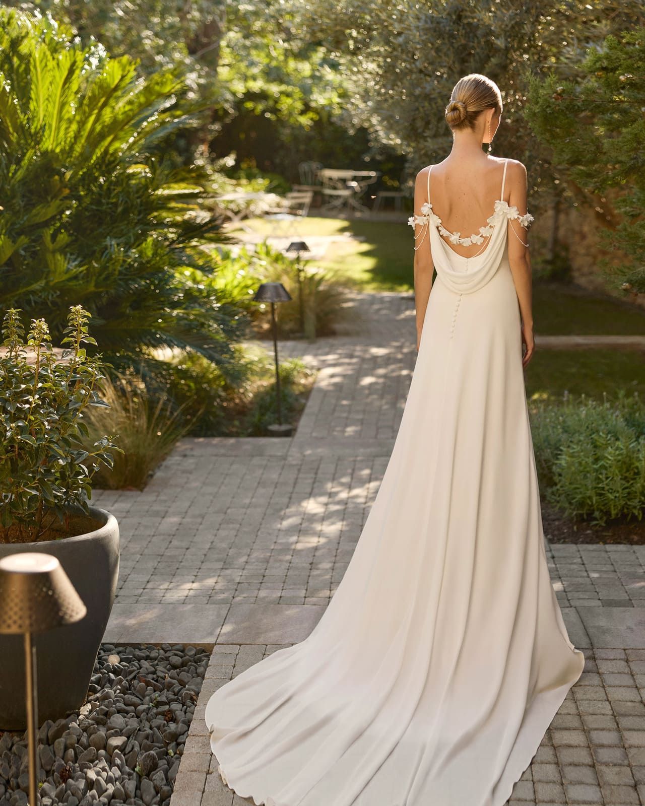 Novia con un largo vestido blanco caminando por un sendero de jardín, vista desde atrás.