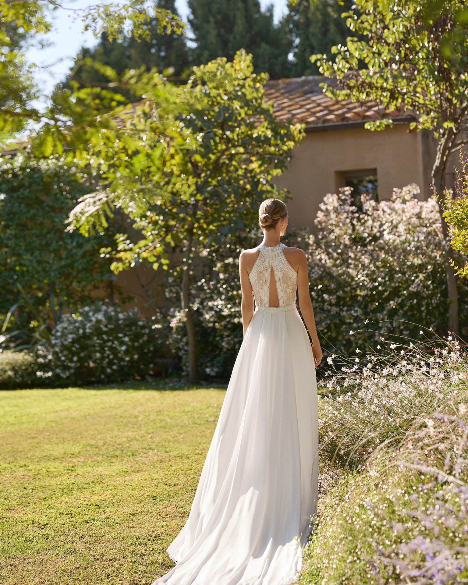 Mujer con un vestido blanco sin espalda de pie en un jardín soleado con árboles y flores.