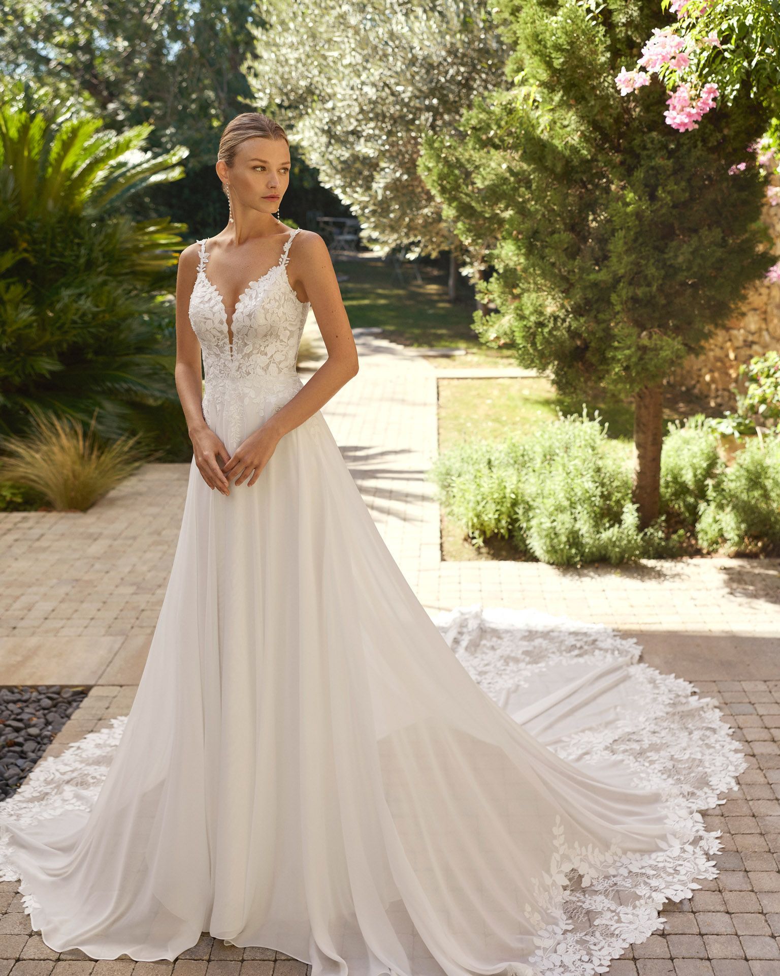 Novia con un vestido de novia de encaje blanco de pie en un sendero soleado de un jardín, con una larga cola.