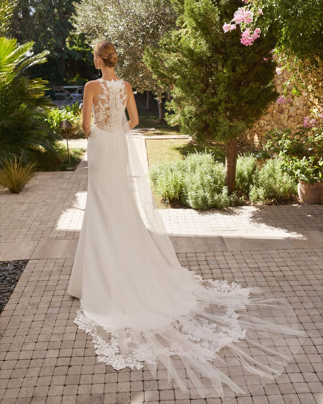 Novia con un vestido blanco de encaje con una larga cola caminando por un sendero del jardín.
