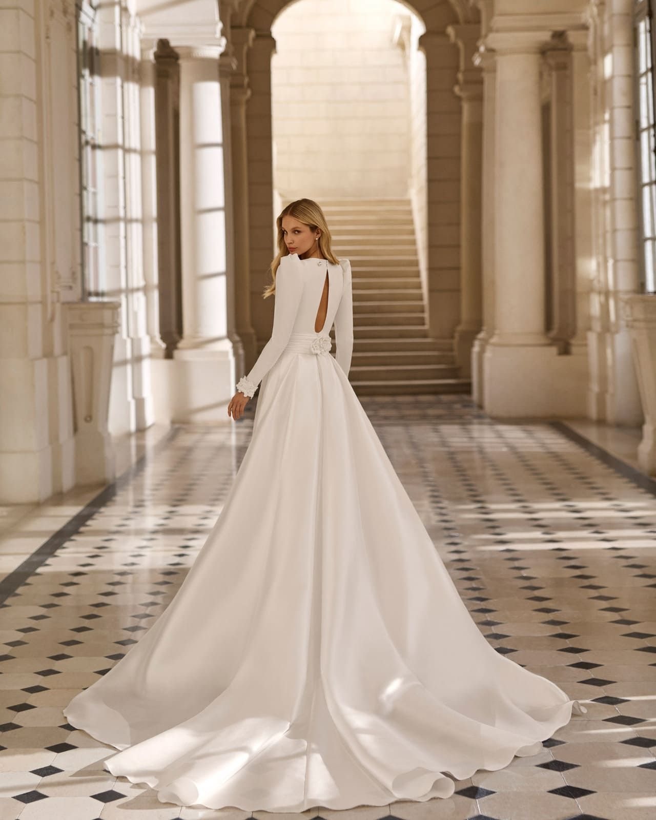 Mujer con un vaporoso vestido blanco de pie en un gran salón de mármol con escaleras a sus espaldas.