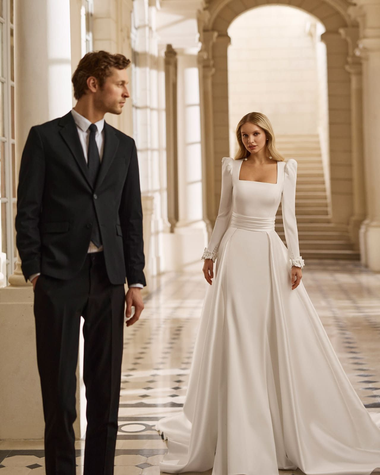 Pareja vestida con traje negro formal y vestido blanco de pie en un gran salón arqueado.