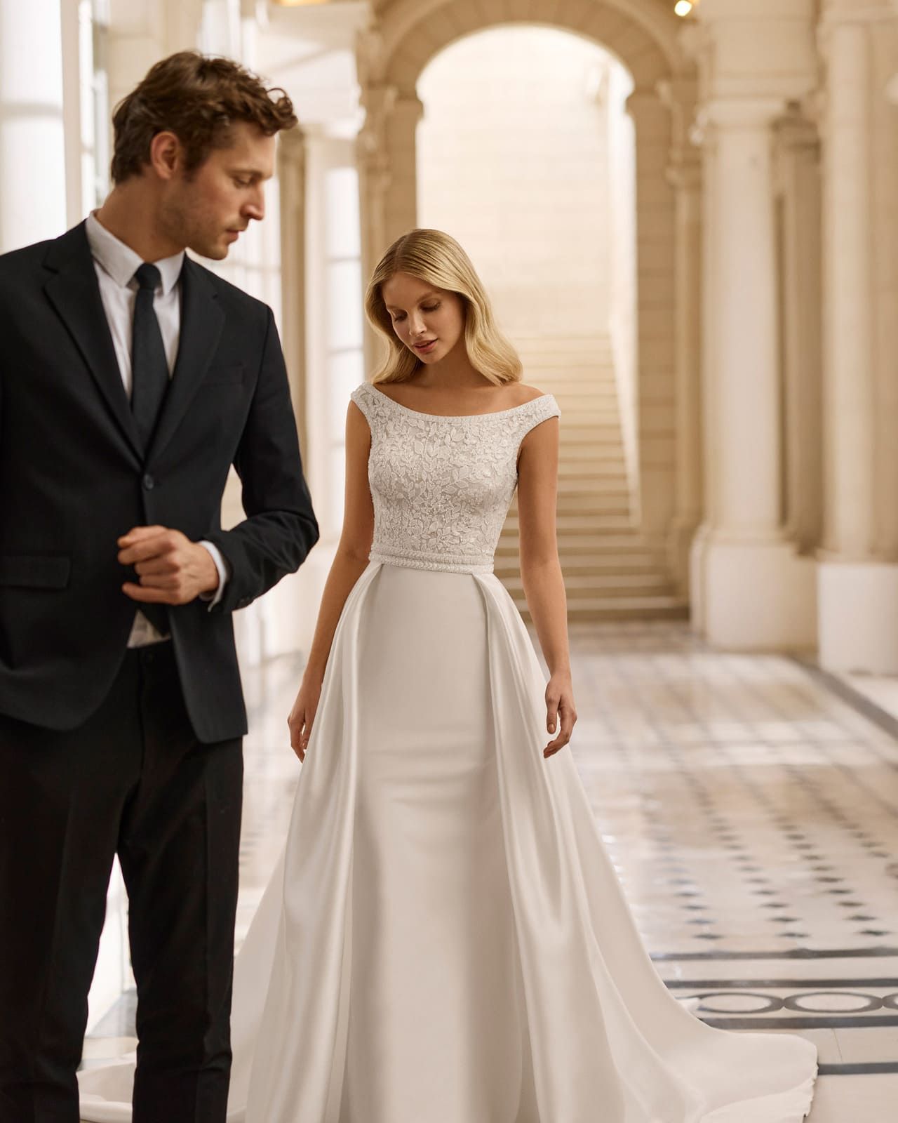La novia, con un vestido blanco de hombros descubiertos, y el novio, con un traje oscuro, caminan por un gran salón.