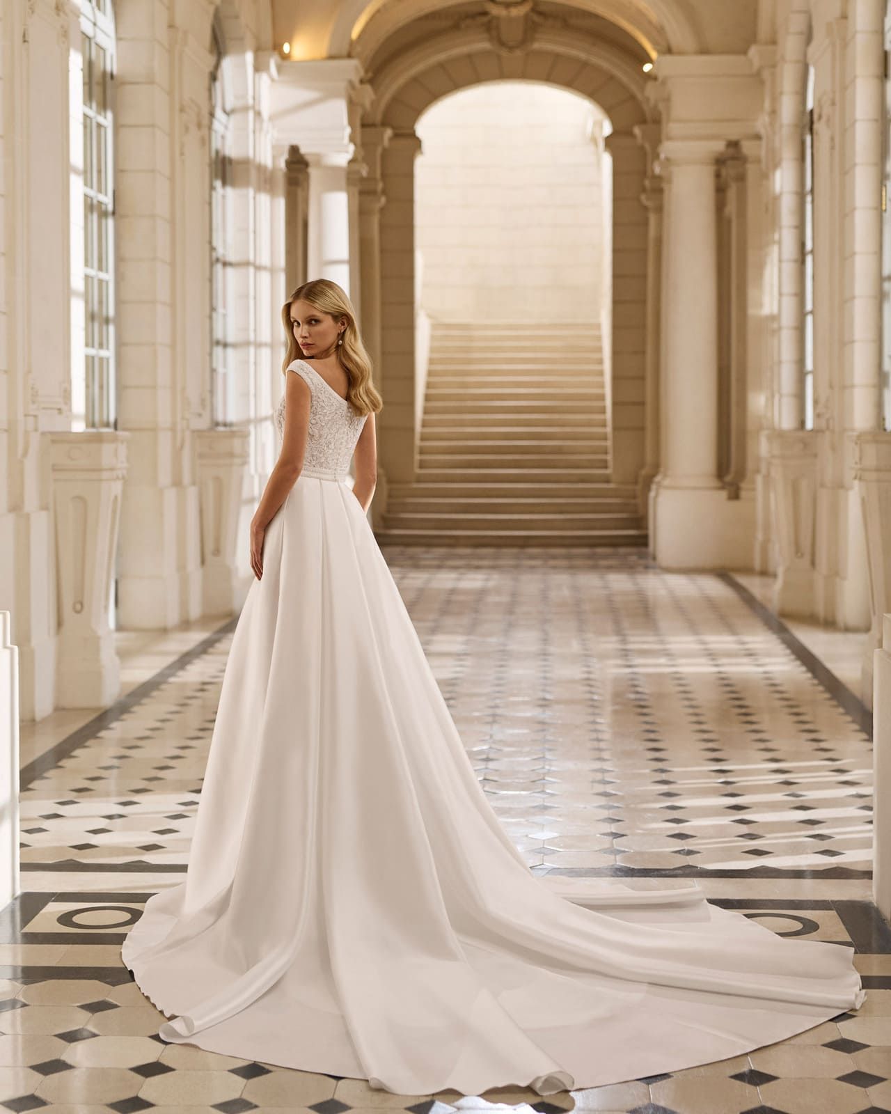 Mujer con vestido blanco de pie en un gran vestíbulo con una majestuosa escalera.