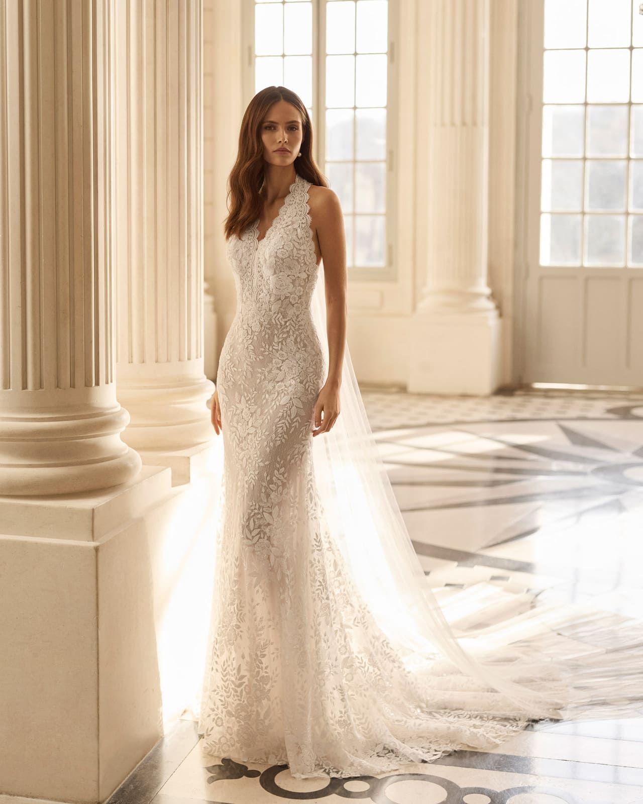 Mujer con un vestido blanco de encaje de pie en un vestíbulo soleado con columnas y grandes ventanales.