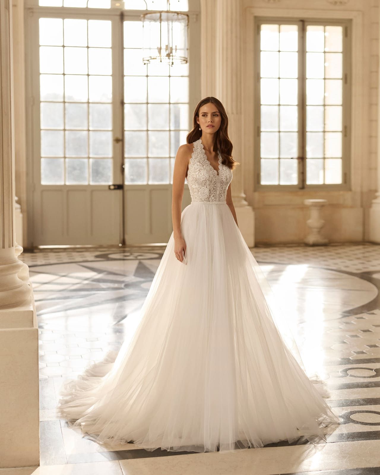 Mujer con un vestido de encaje blanco de pie en un salón de baile iluminado por el sol con grandes ventanales y una falda de tul vaporosa.