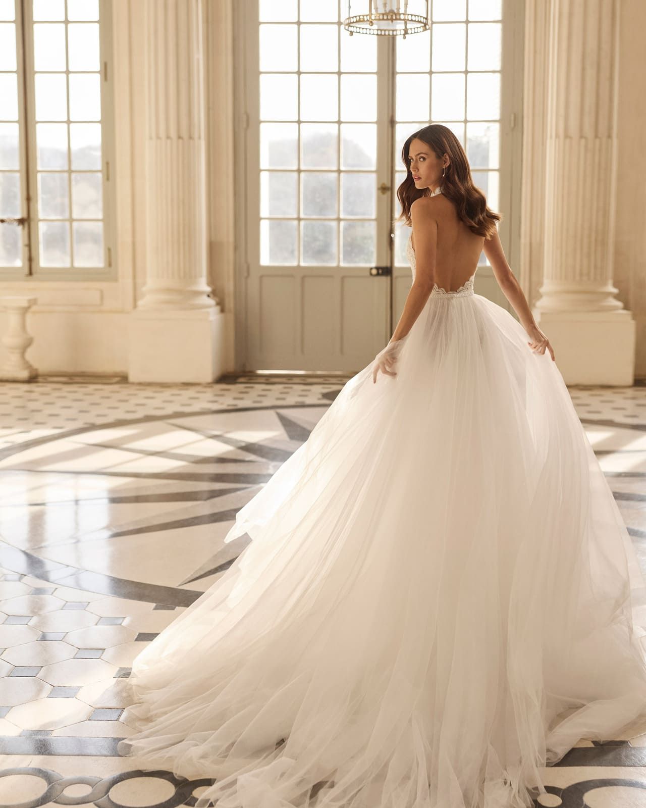 Mujer con un vaporoso vestido blanco en un gran salón con altos ventanales y suelos de mármol.
