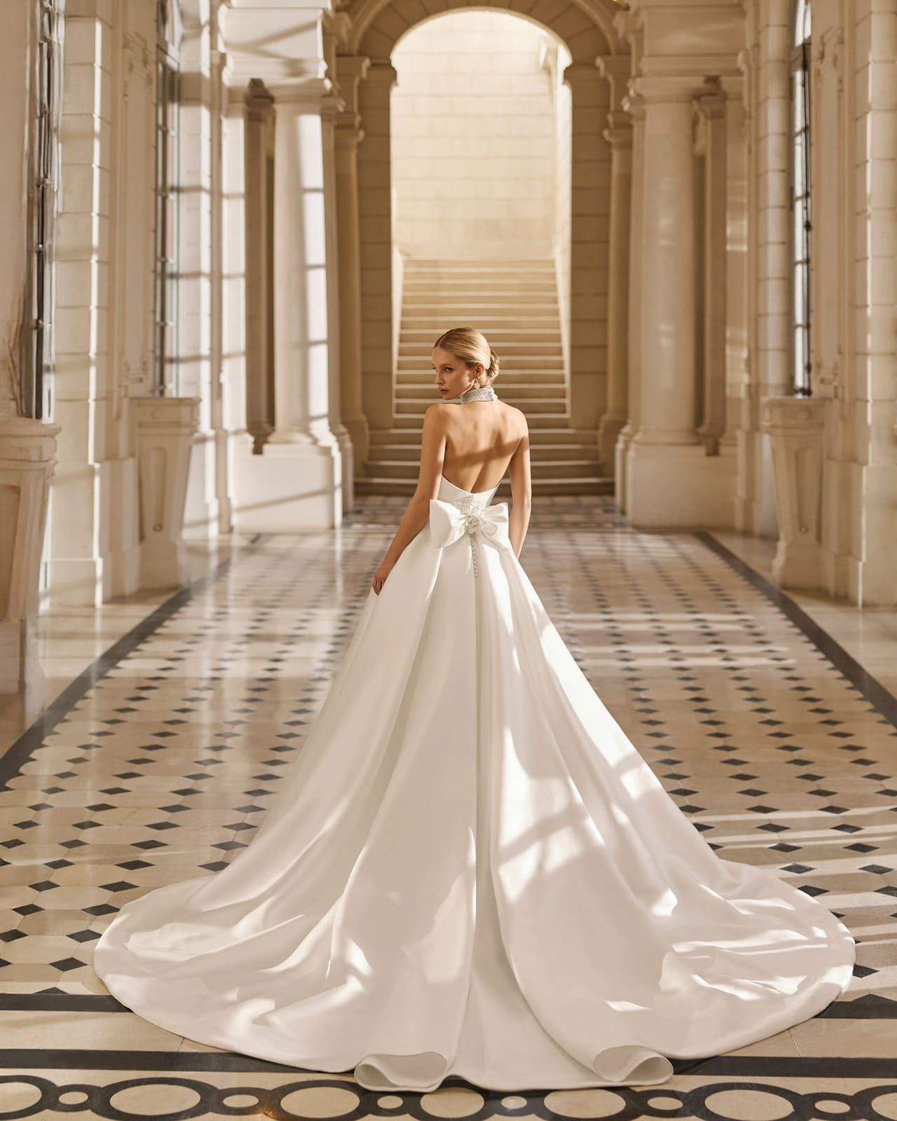 Novia con un vestido blanco de larga cola caminando por un salón de mármol iluminado por el sol.