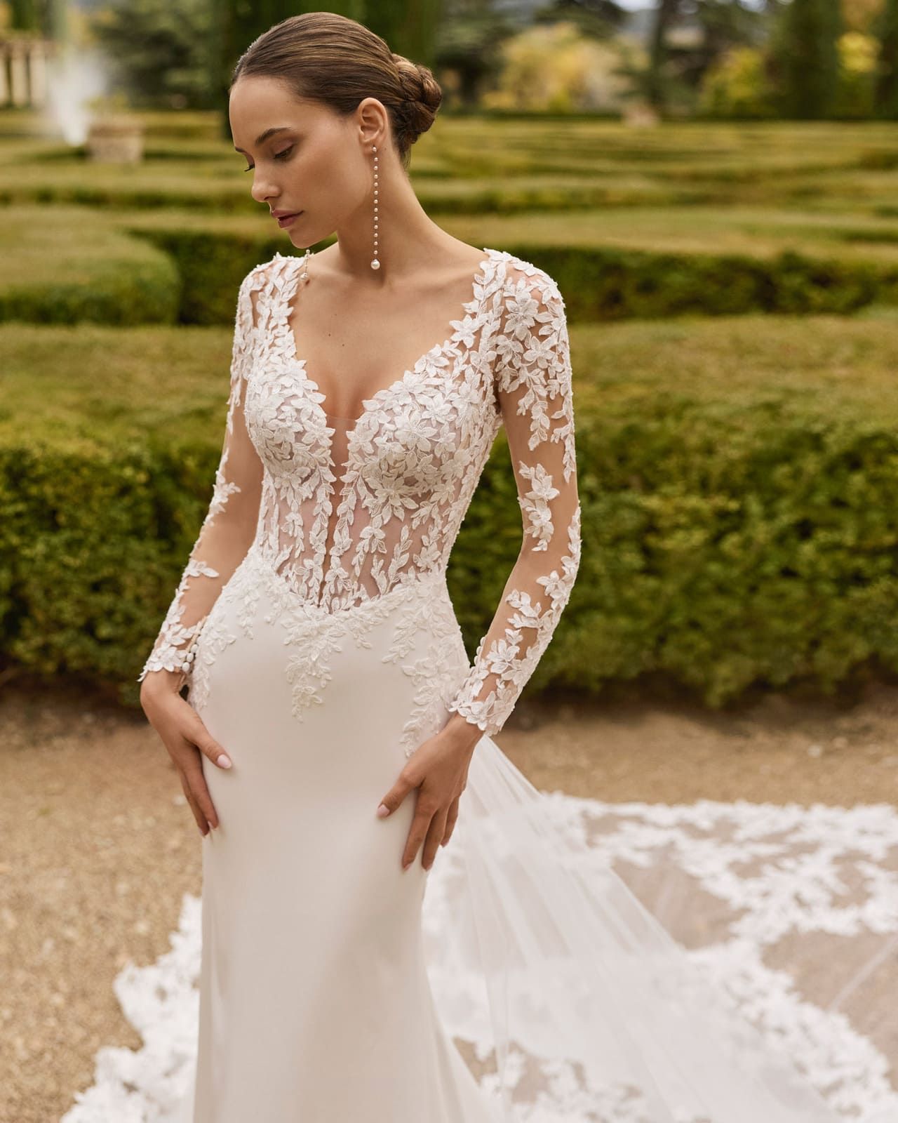 Novia con un vestido de novia de encaje de manga larga de pie en un jardín con una larga cola.