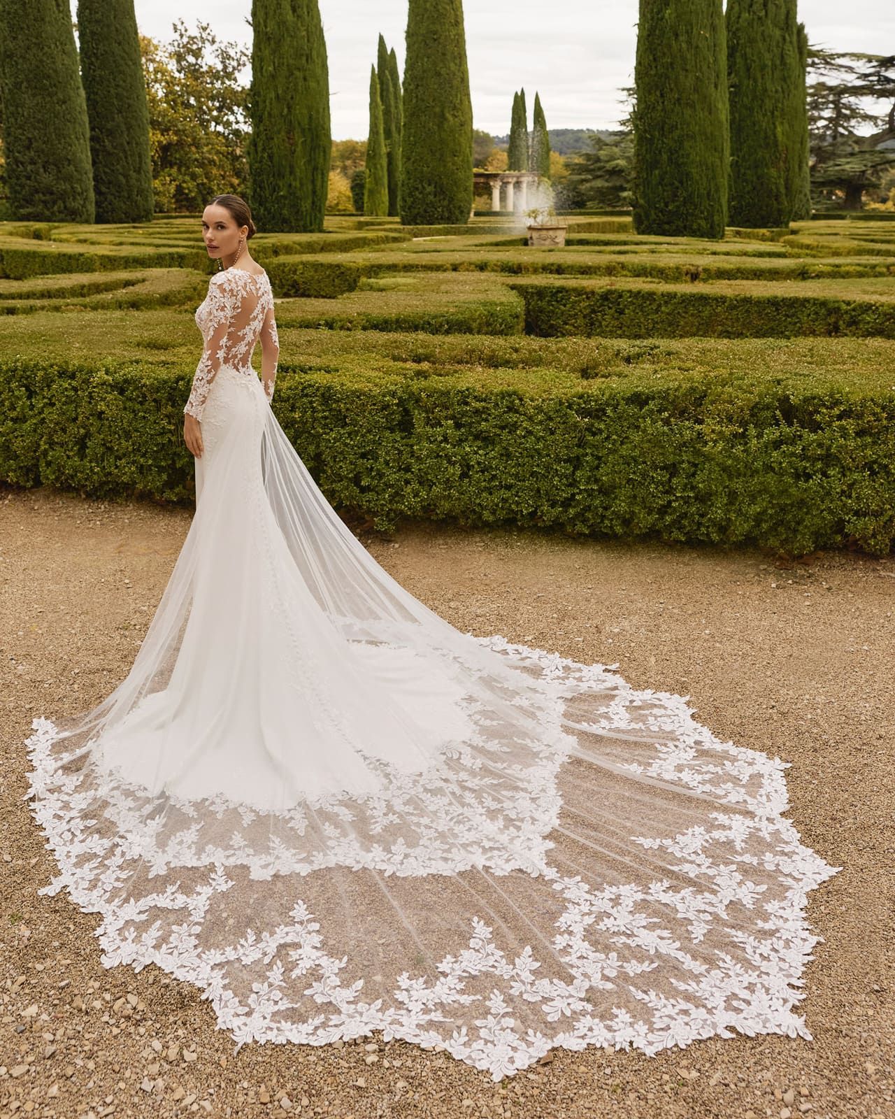 Novia con un vestido blanco de encaje con una larga cola, de pie en un jardín formal con setos y fuentes.