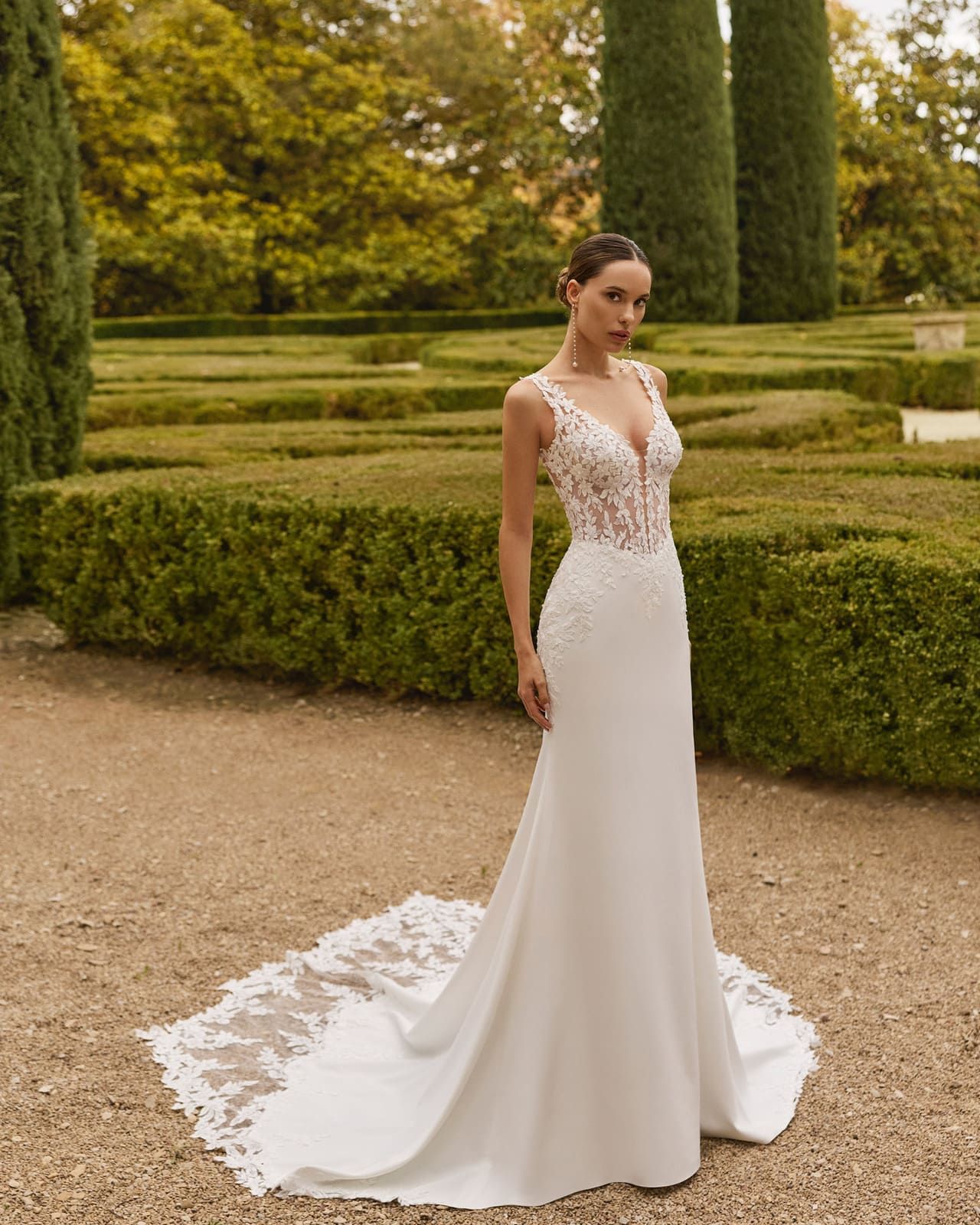 Mujer con un vestido de novia de encaje blanco, de pie en un sendero de jardín bien cuidado, con una larga cola.