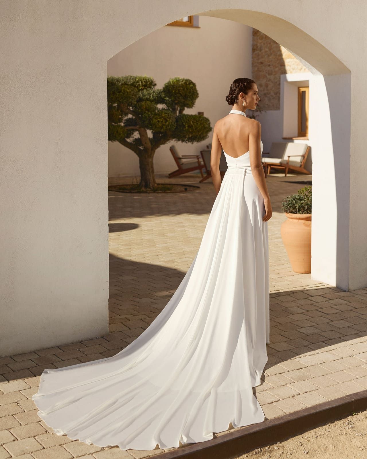 Mujer con un vaporoso vestido blanco sin espalda de pie bajo un arco en un patio soleado.