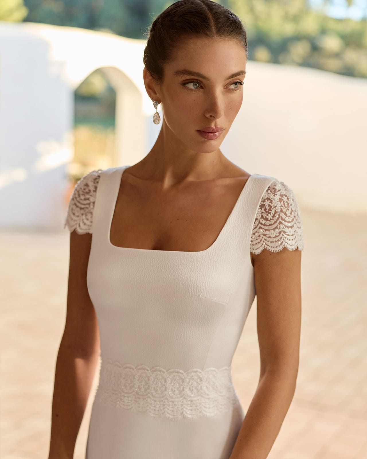 Mujer con un vestido blanco de encaje de pie al aire libre bajo la suave luz del sol, mirando hacia un lado.