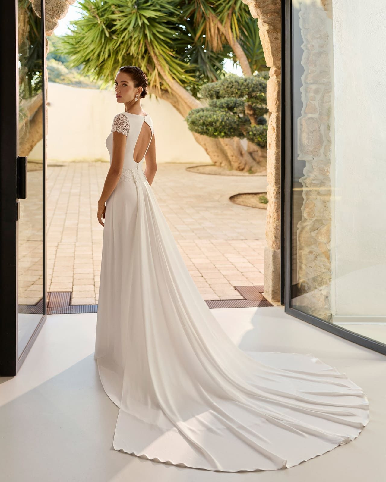 Novia con vestido blanco de pie en el umbral de una puerta con una larga cola en un patio soleado.