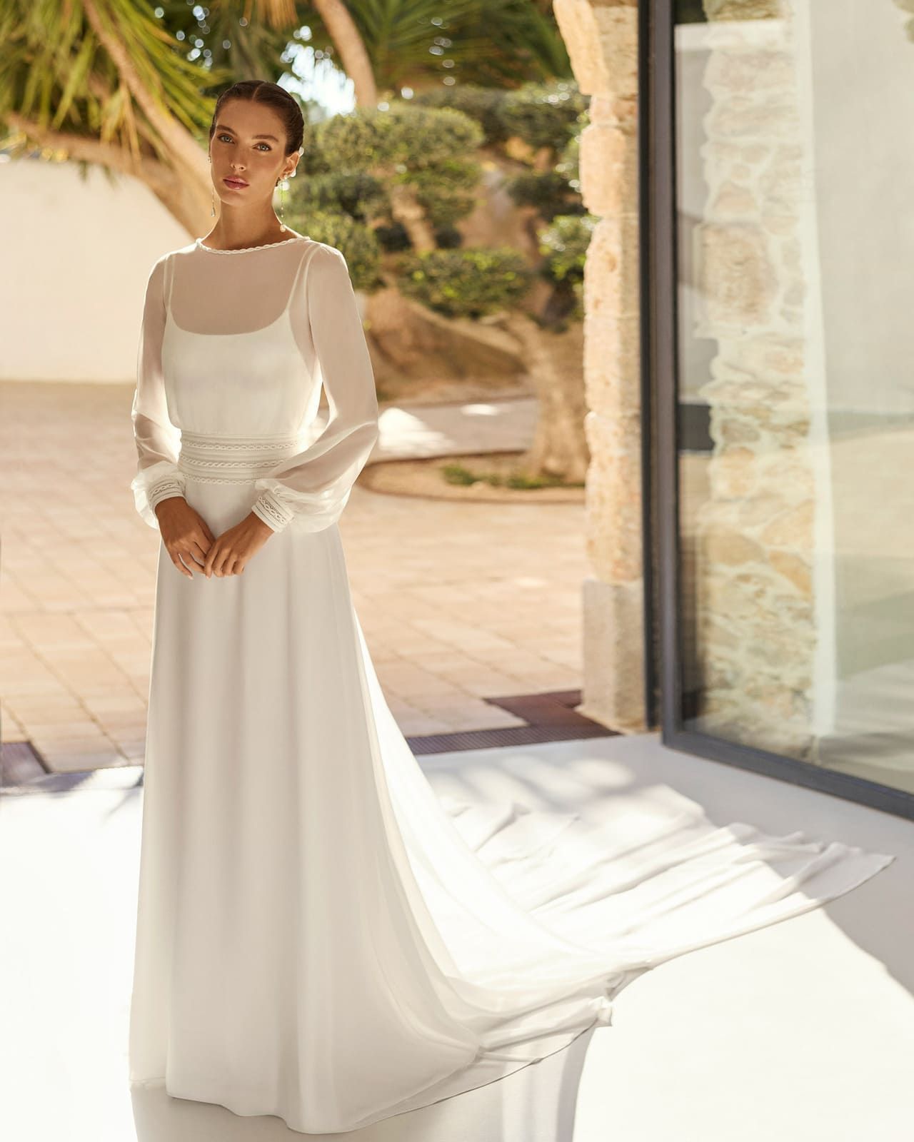 Novia con vestido de novia blanco de pie en el interior de una casa, cerca de una puerta iluminada por el sol.