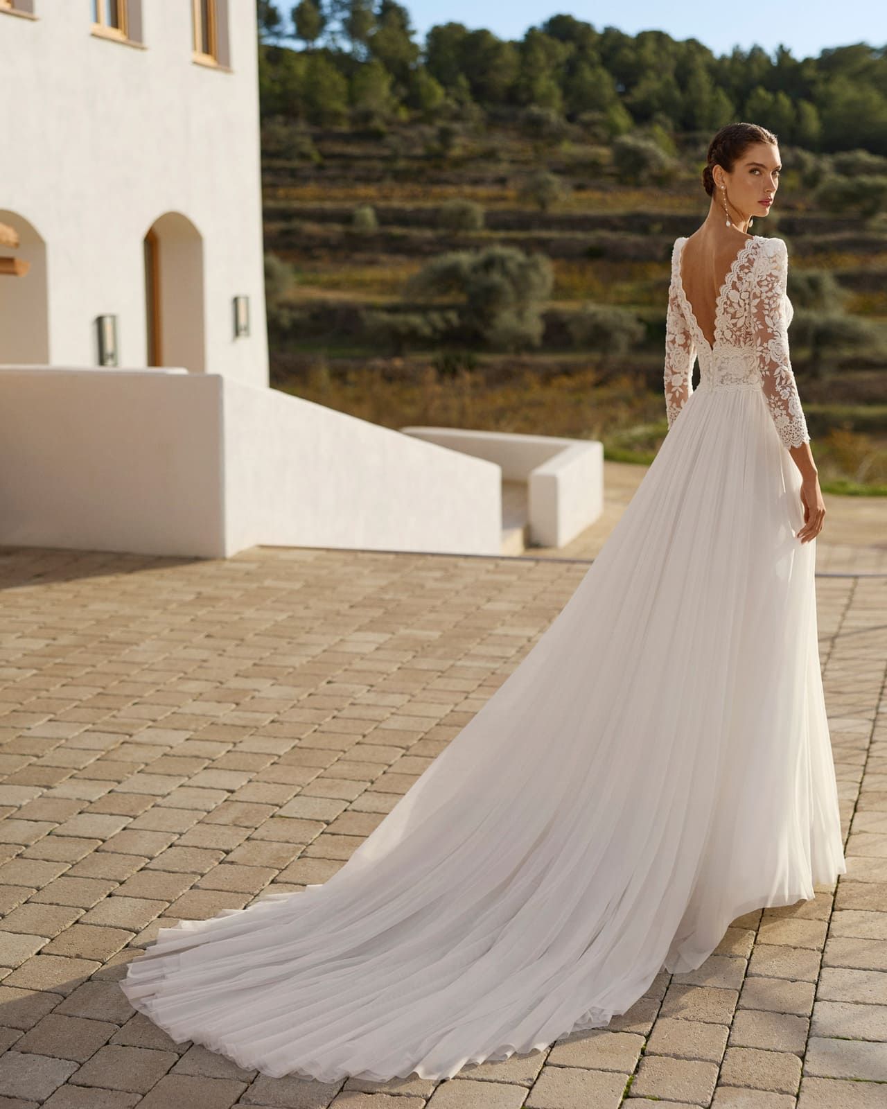 Mujer con un vestido de novia blanco de encaje con una larga cola de pie en una terraza soleada.