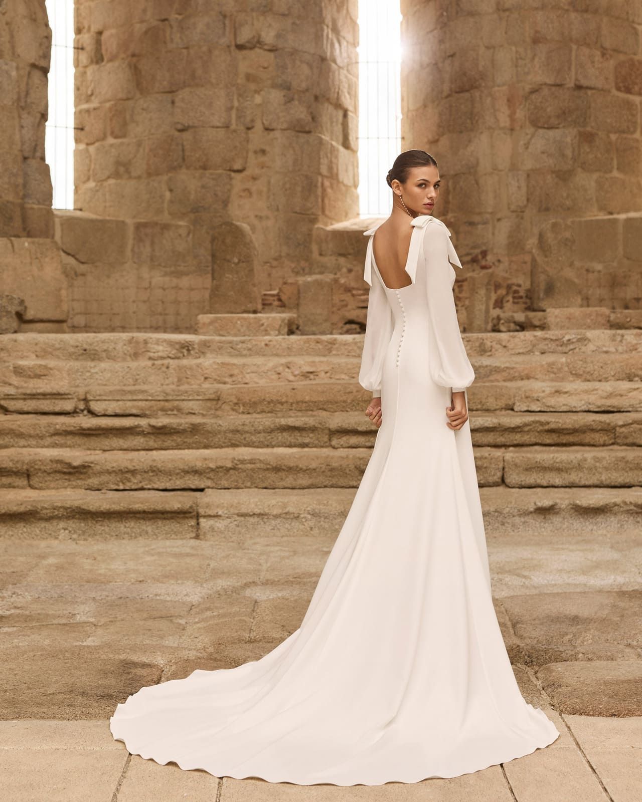 Mujer con un vestido blanco vaporoso de pie sobre escalones de piedra frente a columnas antiguas.