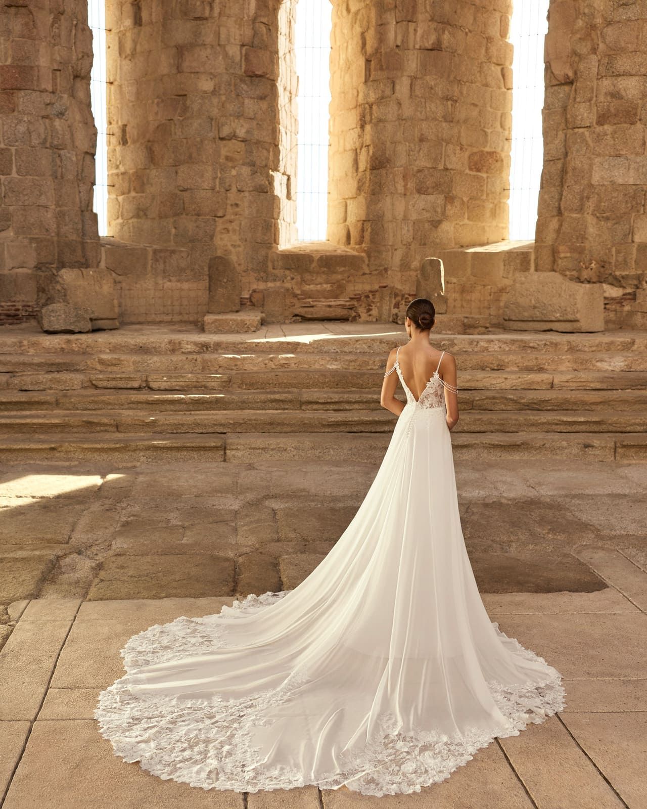 Mujer con un vestido de novia blanco con una larga cola de pie en una sala de ruinas de piedra.