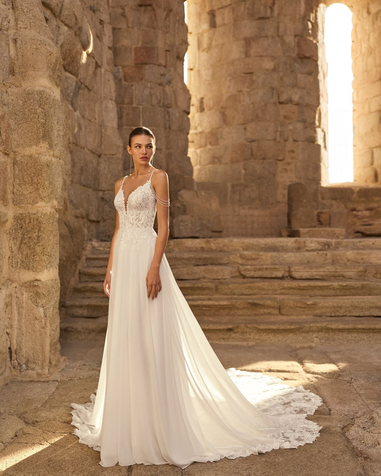 Mujer con un vestido de novia blanco de pie entre ruinas de piedra, con una larga cola y bañada por la brillante luz del sol.