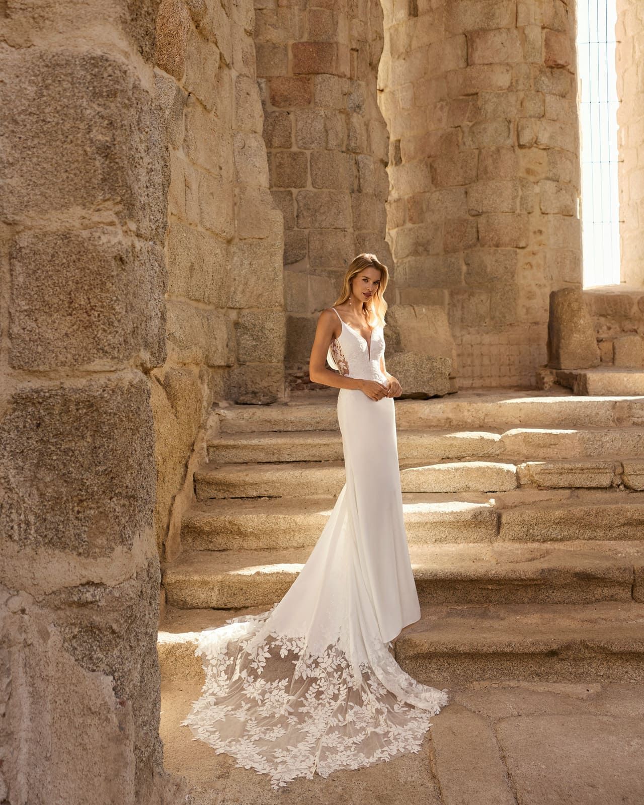Mujer con vestido de encaje blanco de pie sobre escalones de piedra en un antiguo entorno pétreo iluminado por el sol.