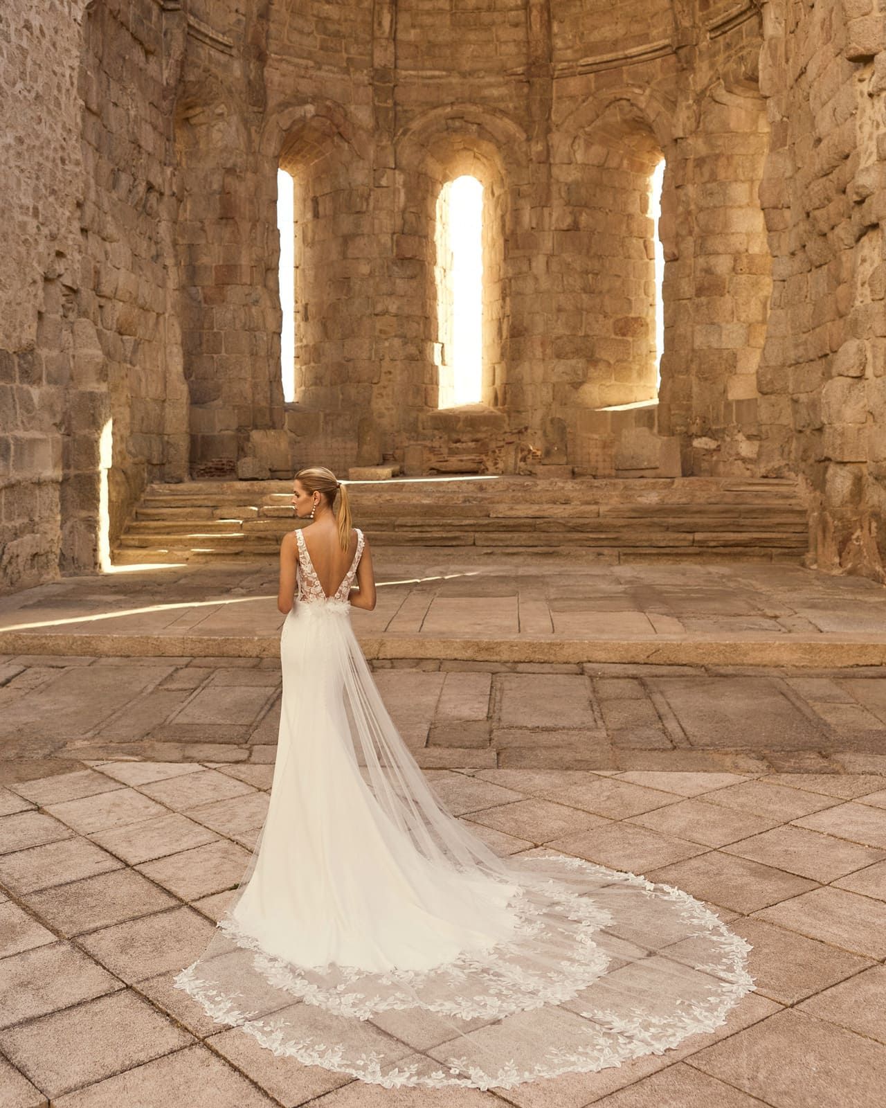 Novia con vestido blanco de larga cola de pie en un salón de piedra iluminado por el sol.