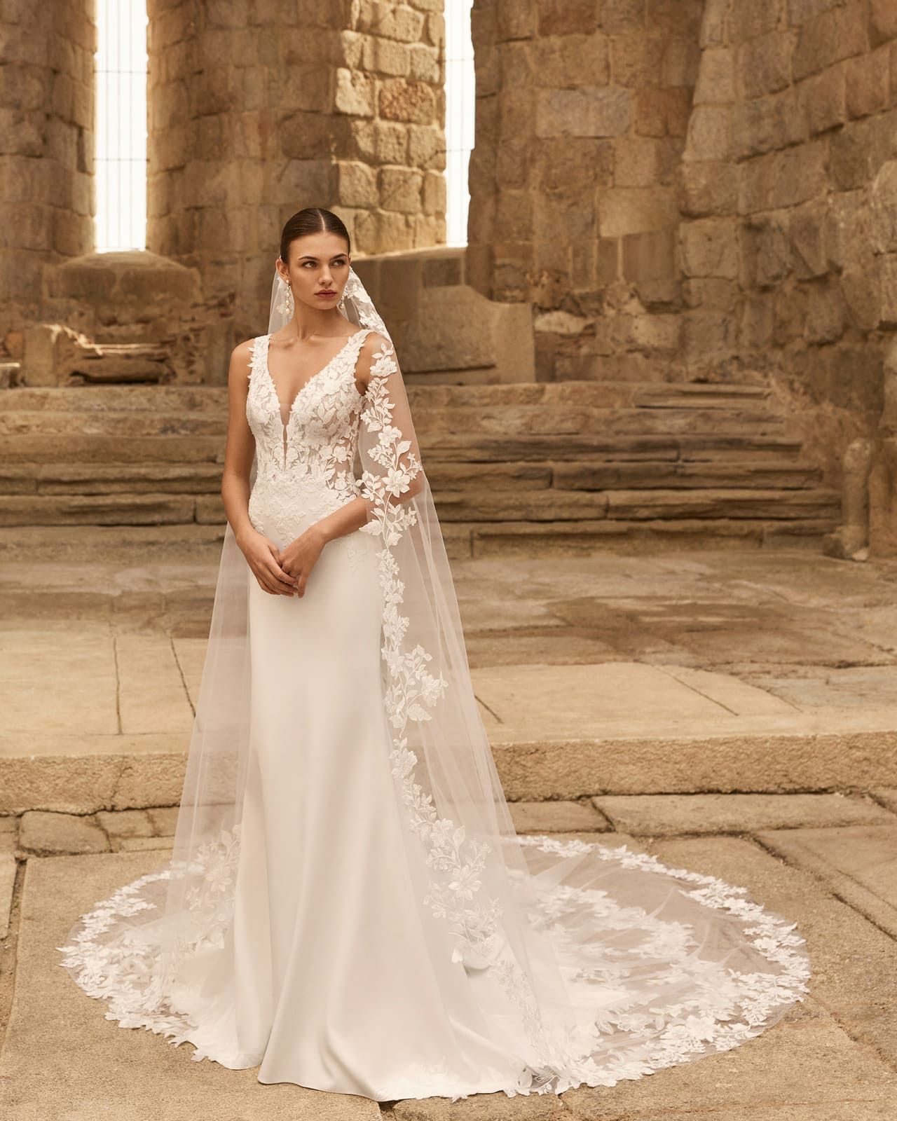 Novia con vestido blanco y velo de encaje de pie en un entorno de ruinas de piedra.