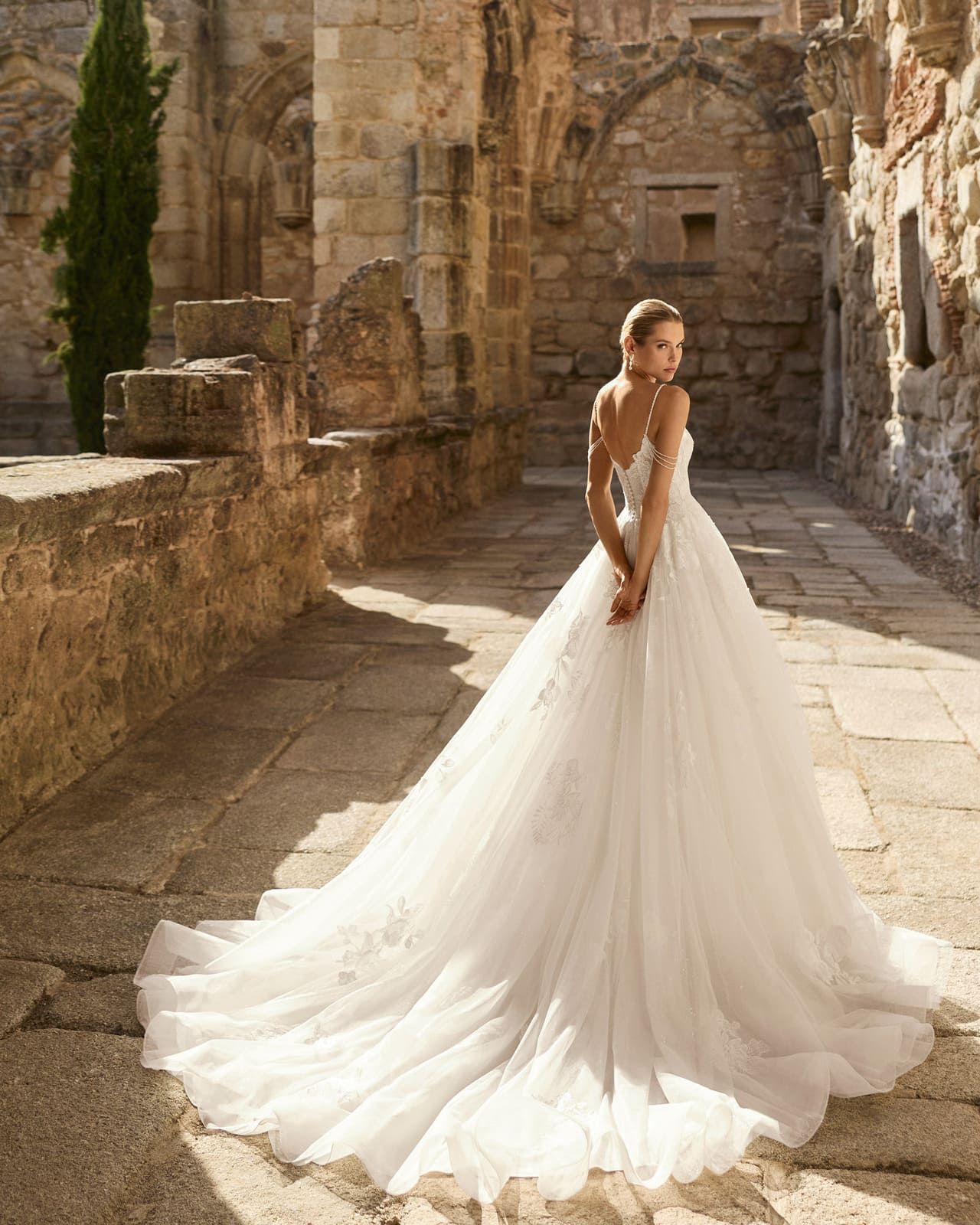 Novia con un largo vestido blanco de pie en un patio de piedra bañado por el sol.