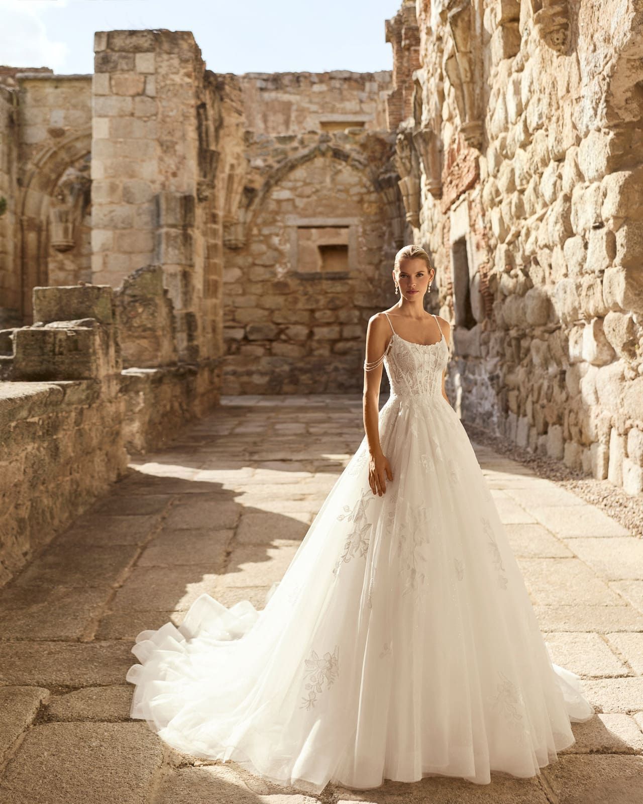 Novia con un vestido de novia blanco de pie entre ruinas de piedra iluminadas por el sol con una larga cola ondeante