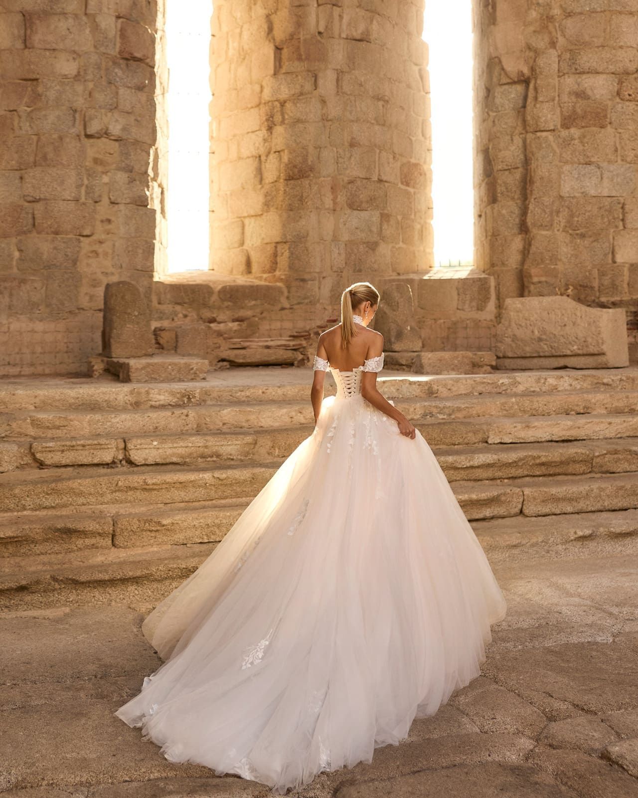 Novia con un vaporoso vestido blanco de pie sobre escalones de piedra en una antigua ruina bañada por el sol.
