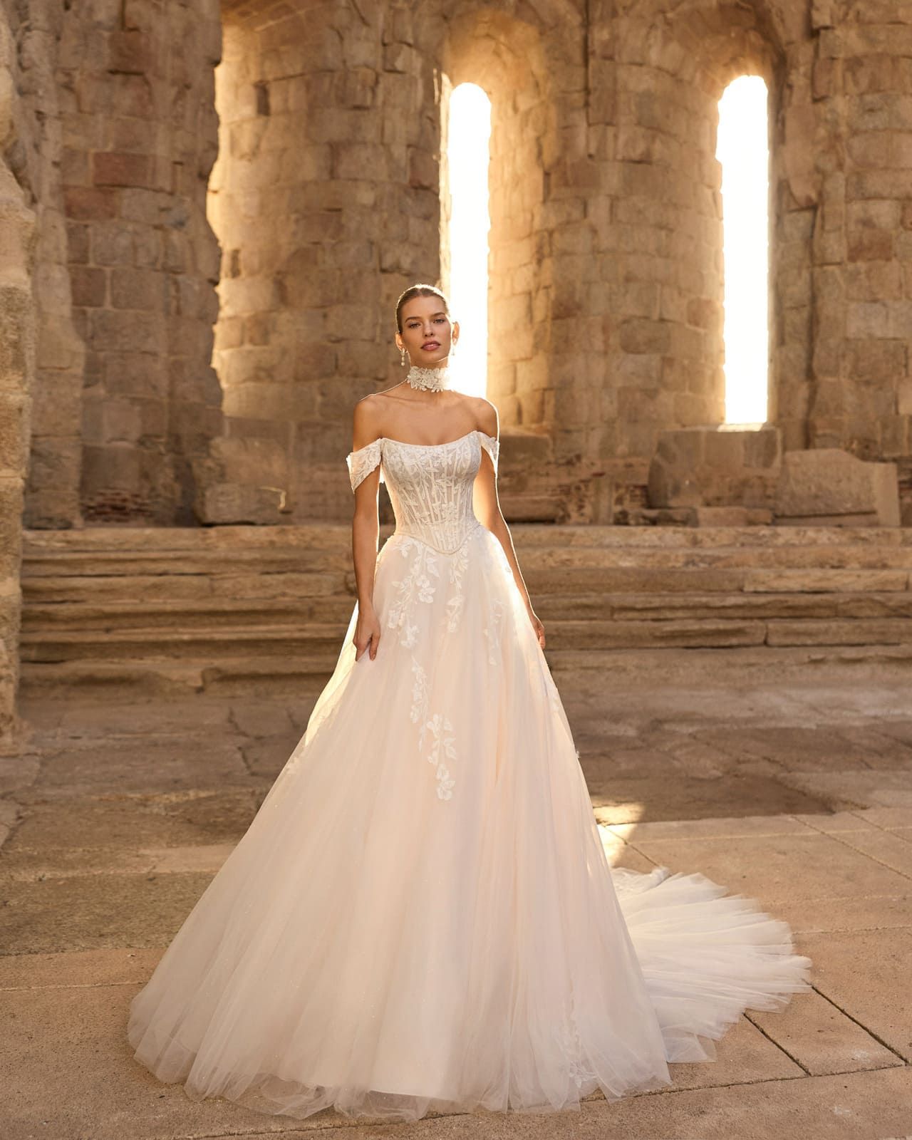 Novia con un vestido de novia blanco sin hombros de pie en un salón de piedra con altas ventanas arqueadas.