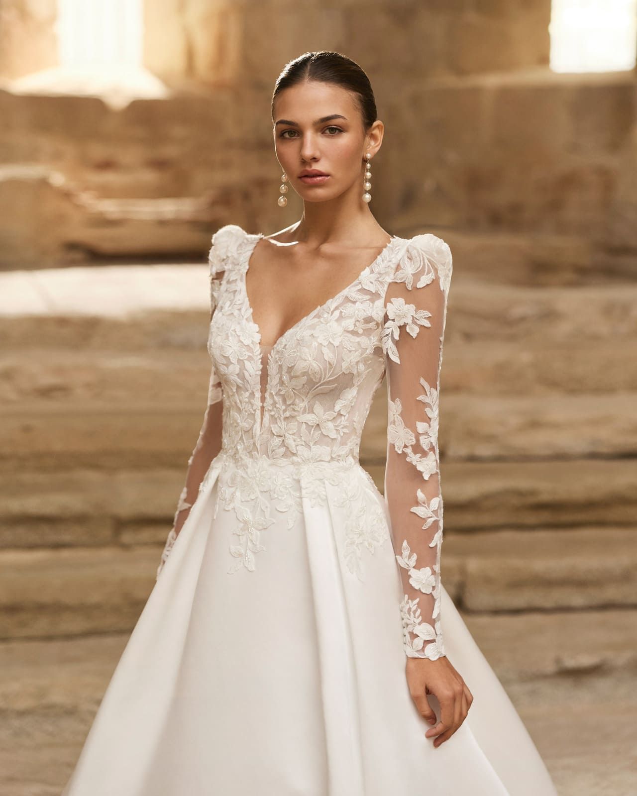 Mujer con un vestido de novia de encaje blanco de pie sobre escalones de piedra en un entorno de tonos cálidos.