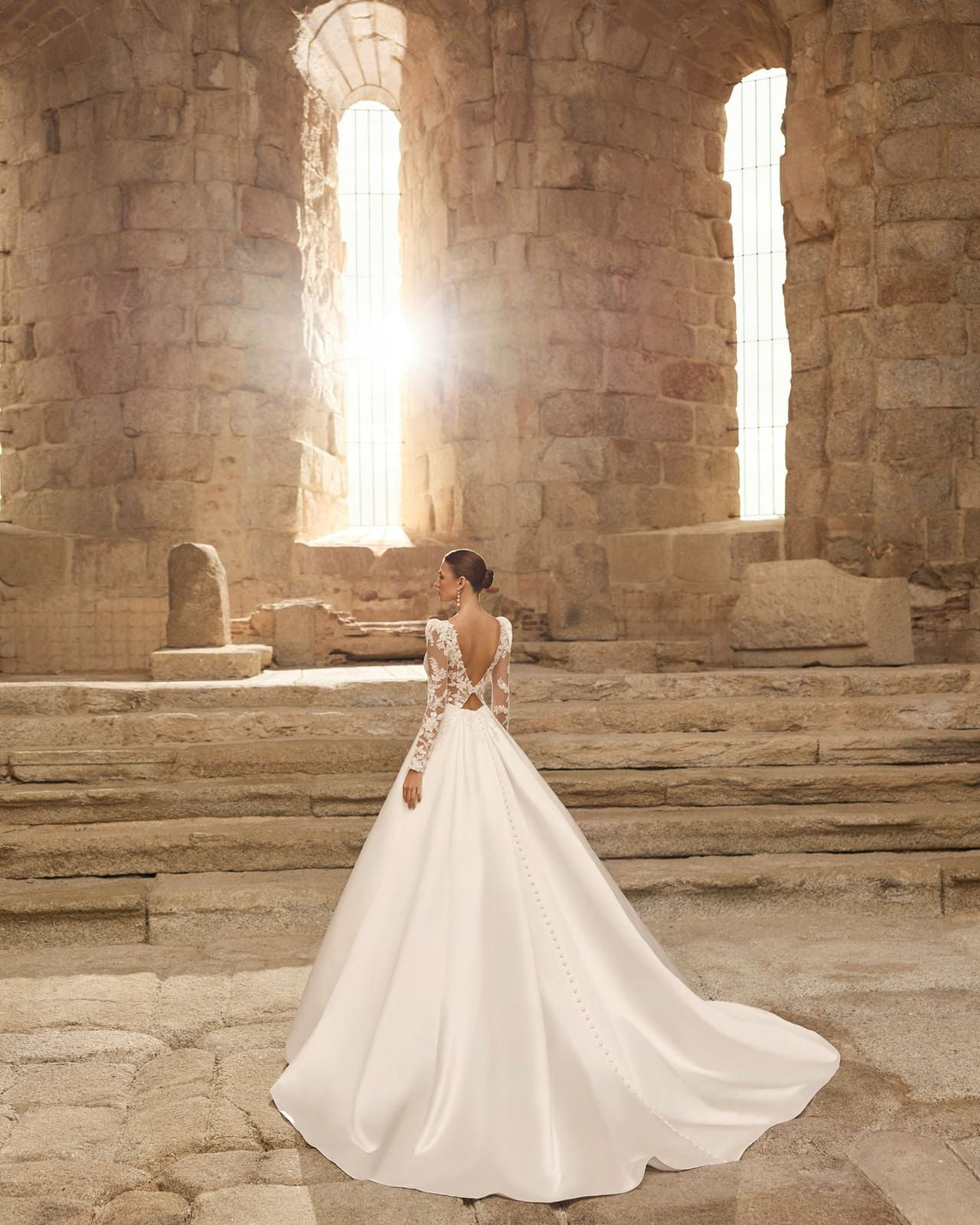 Novia con vestido blanco de pie sobre escalones de piedra dentro de un salón de piedra bañado por el sol.