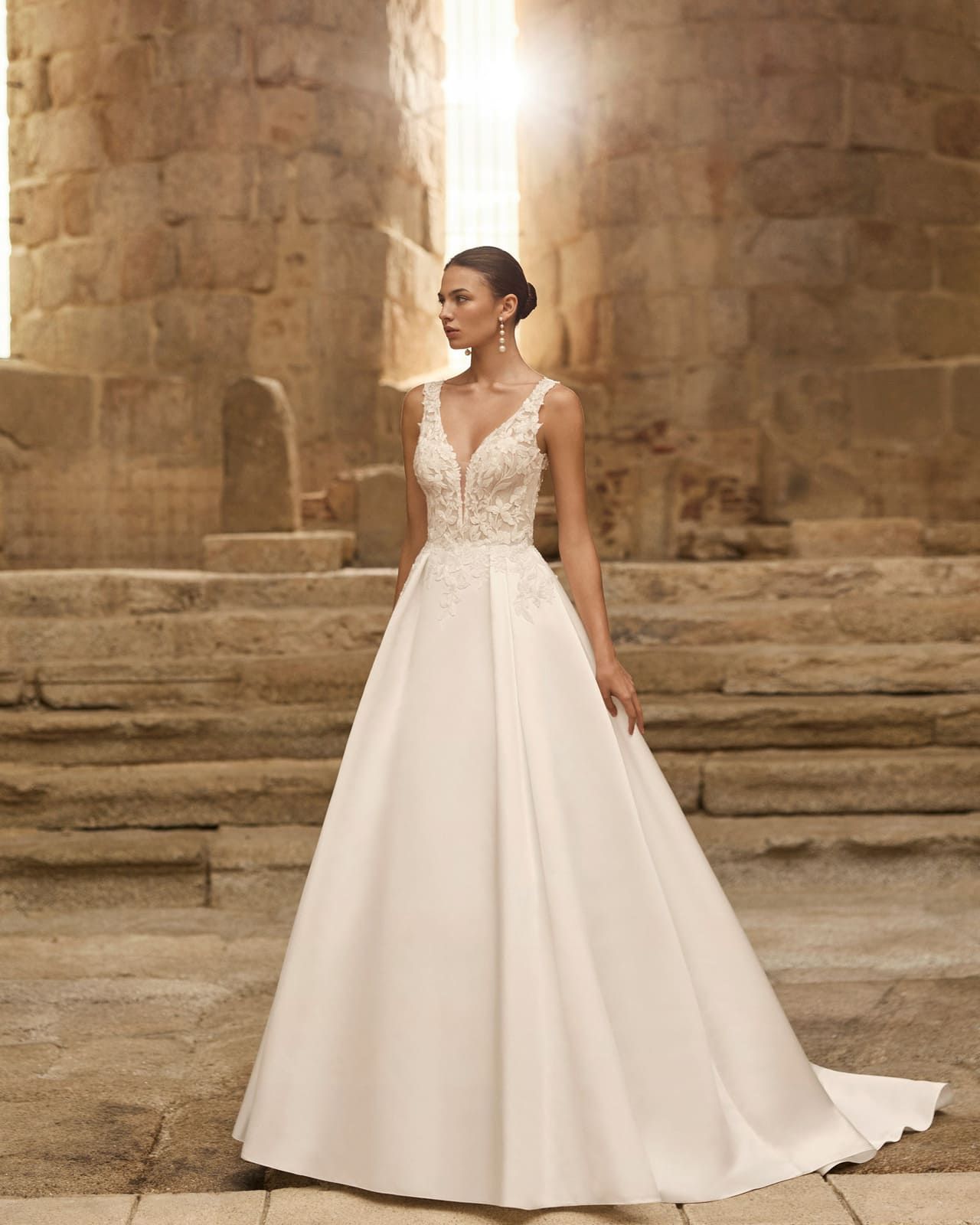 Mujer con vestido de novia blanco de pie sobre escalones de piedra en un gran salón iluminado con luz cálida.