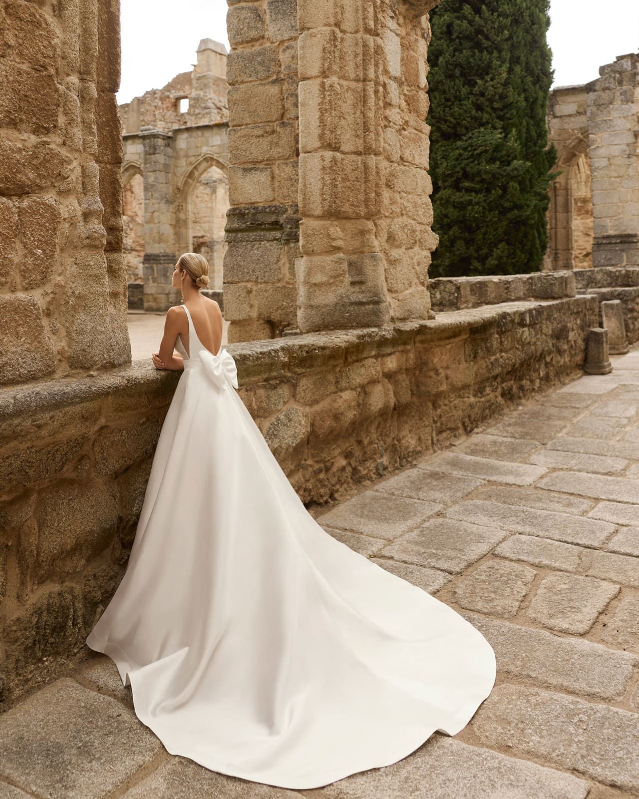 Novia con un vestido blanco de pie junto a antiguas ruinas de piedra, con una larga cola sobre un camino empedrado.