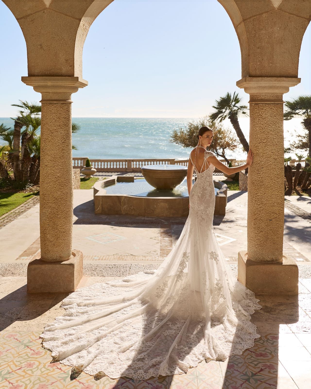 Novia con vestido blanco de pie bajo arcos de piedra con vistas al mar al atardecer.