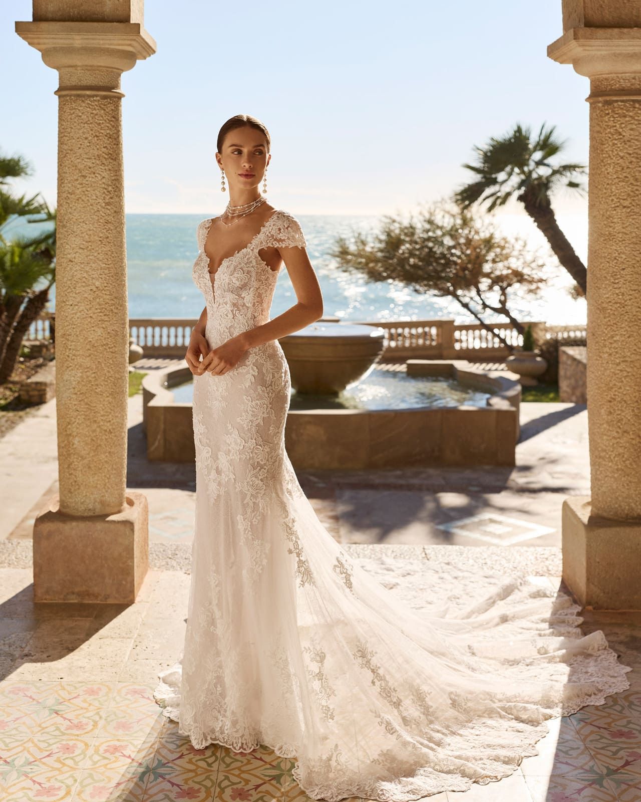 Novia con vestido de novia de encaje de pie en una terraza junto al mar con el océano y palmeras detrás de ella.