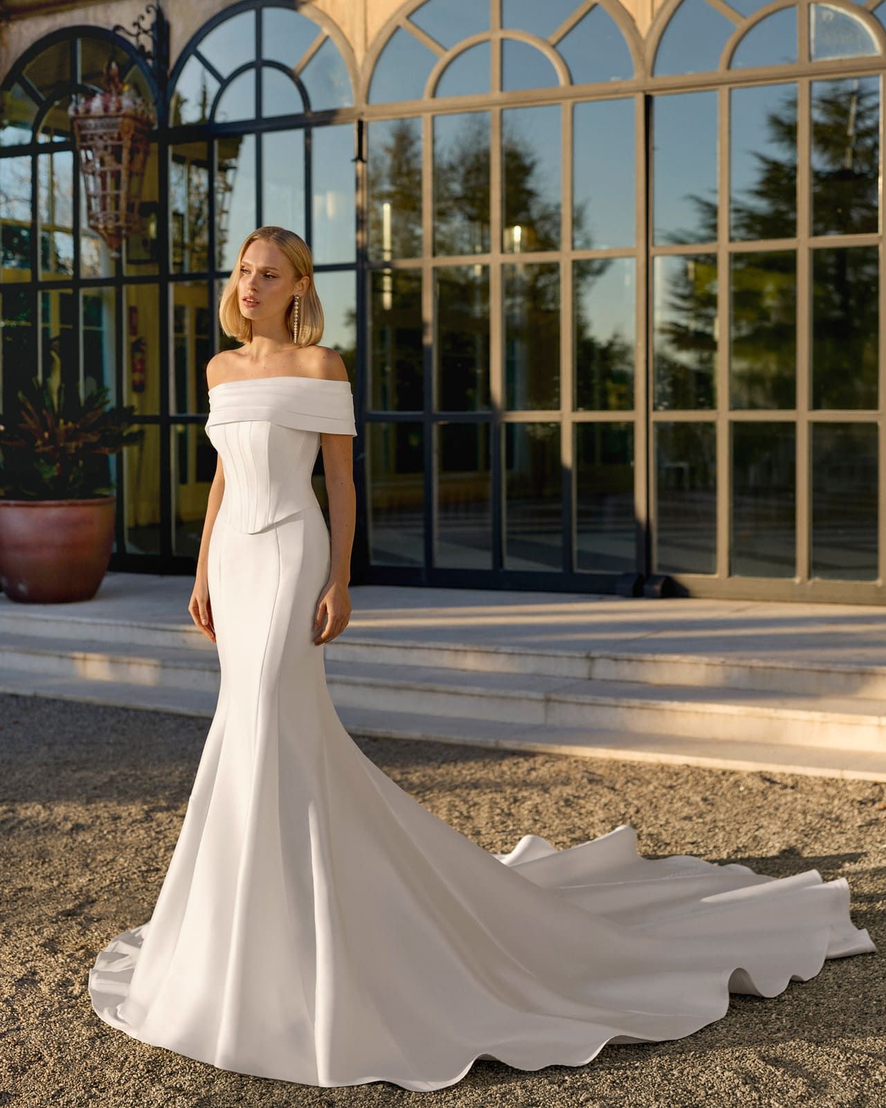 Mujer con un vestido blanco sin hombros de pie al aire libre frente a unas puertas de cristal arqueadas.